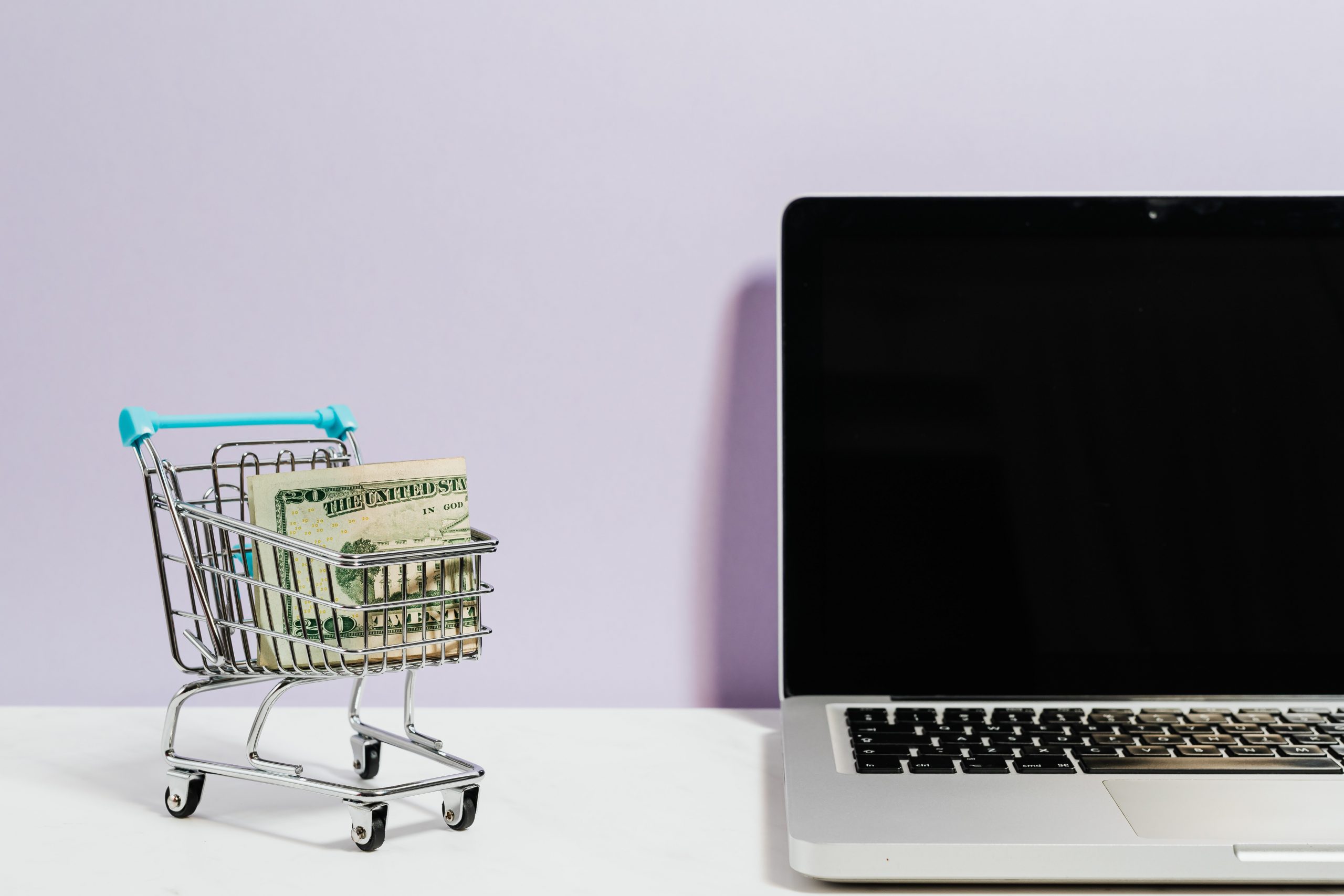 Laptop on a desk next to a tiny shopping cart with cash in it